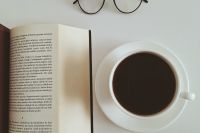 Na zdjęciu na białym stole kawa, książka i okulary.